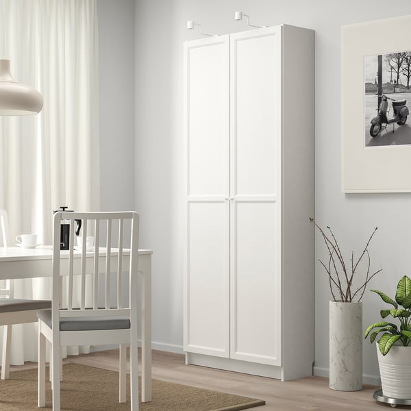 A modern, white BILLY bookcase stands tall, near a dining table and chair, offering ample storage. Its sleek design fits well in the bright room.