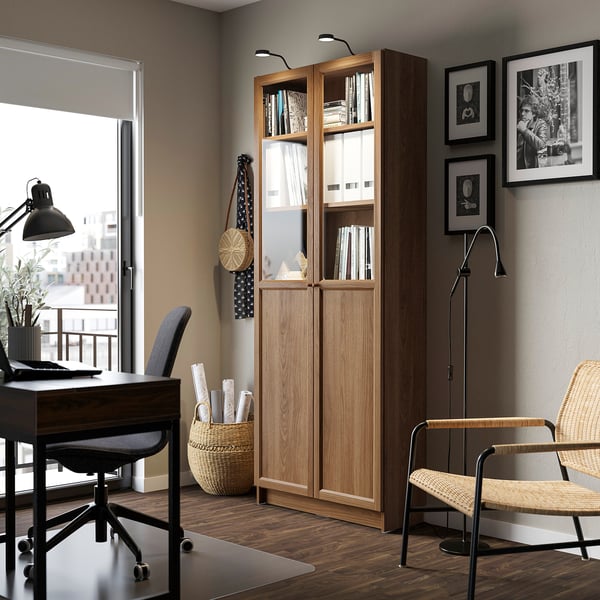 Bookcase with glass doors near window, books, decor, desk, lamp, pictures. Well-lit, organised.