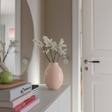 Vase named BLODBJÖRK, light pink, made of stoneware, sits on a shelf. Its small size contrasts with stacked books and a door.