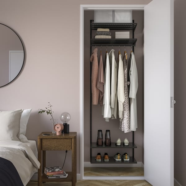BOAXEL closet storage with clothes and accessories. Steel, wall-mounted, dark grey. Nearby bedside table with lamp, clock, plant.