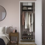 BOAXEL closet storage with clothes and accessories. Steel, wall-mounted, dark grey. Nearby bedside table with lamp, clock, plant.