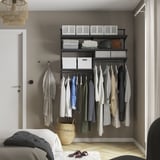 BOAXEL closet with black shelves and hanging rail, holding clothes and accessories. Grey wall, white door, basket, slippers on floor.