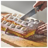 A person is cutting a rectangular cake with white toppings on a wooden board using a BRILJERA knife.