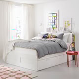 Modern white bed with grey bedding, colorful pillows, and red side table; bright, minimalist bedroom.