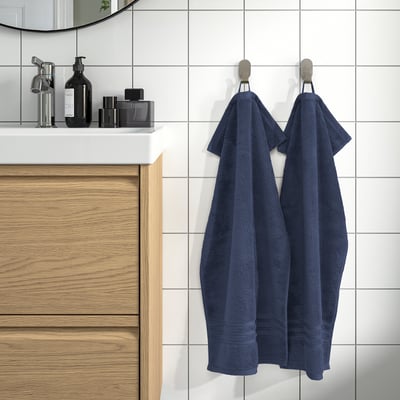 Tidy bathroom with wooden vanity. Large navy blue BROKGLIM towels hang neatly on wall hooks. White tiles contrast with warm wooden cabinets.