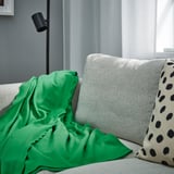 A cozy green DAGGKÅPOR throw blanket draped over a large grey couch, complementing a spotted pillow.