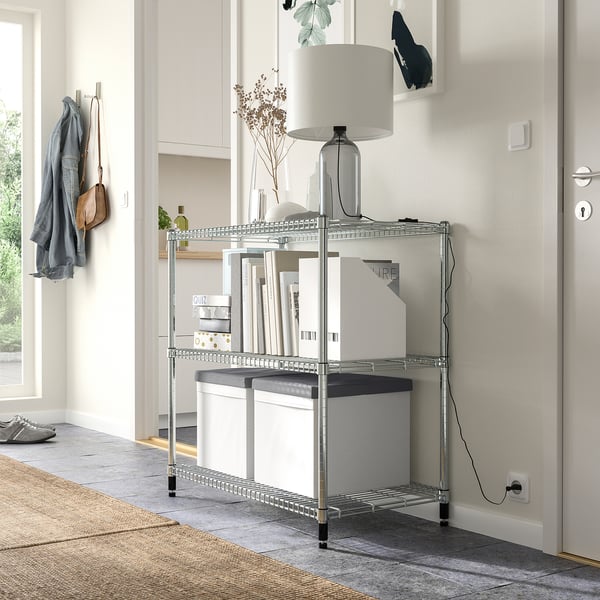 A modern entryway with a metallic 3-tier storage shelf holding white boxes, a lamp, and books. A grey jacket and bag hang nearby.