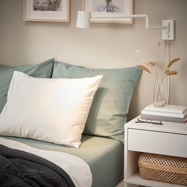 Cozy bedroom corner with DVALA grey-green pillow, light blue sheets, white pillows, books, dried flowers, and wall lamp.