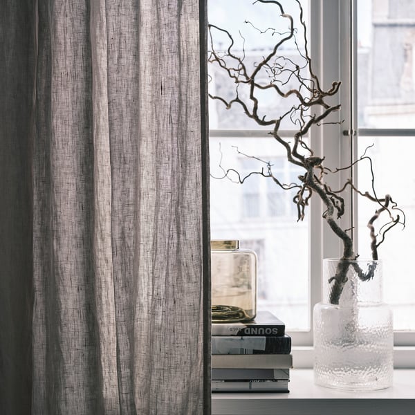 Gray curtains beside window, filtered daylight, vase with branches, stack of books.