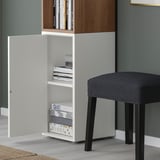 White EKET storage unit with open door, games, and books; gray upholstered stool beside it.