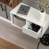 White EKET storage unit with two open compartments, one containing a gray felt bin and the other a black drawer with visible books and gloves.