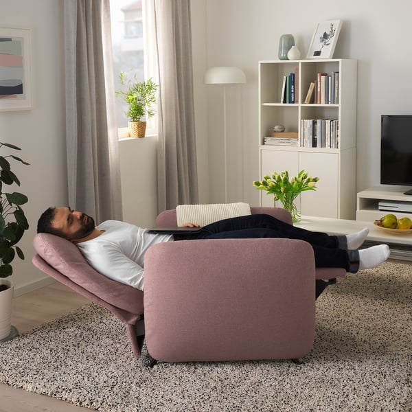 Man relaxes on adjustable pink EKOLSUND recliner in living room. Chair reclines, offering nap comfort. Nearby bookshelf, plants, flowers, and tv.