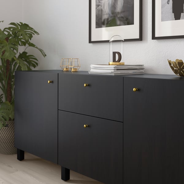 Dark cabinet with gold handles, featuring stacked books and decor items. A large plant is nearby. Framed pictures hang on the wall above.