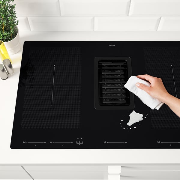 Person cleaning modern black induction hobby with white cloth. Hob has built-in extractor fan, sleek layout for kitchen island.