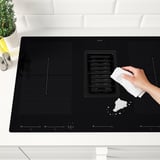 Person cleaning modern black induction hobby with white cloth. Hob has built-in extractor fan, sleek layout for kitchen island.