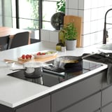 Modern kitchen with island featuring FÖRDELAKTIG induction hob and extractor, sleek black and white design, pots cooking, fresh ingredients nearby.