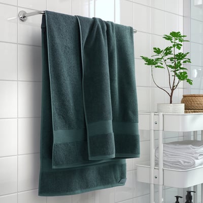 Green FREDRIKSJÖN towels on white rod, tiled wall, storage rack with extra towels and plant.