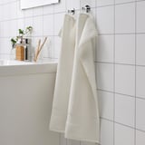 White FREDRIKSJÖN bath towels hang in tiled bathroom, larger than toothbrush holder and vase.