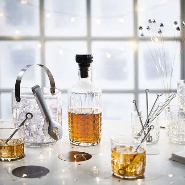 Cocktail setup: clear glass bottle with black cap, ice bucket, tongs, glasses, stirrers on white table.