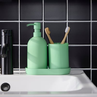 Green soap dispenser and toothbrush holder on white sink. Tray keeps items organized. Against dark tiled wall.