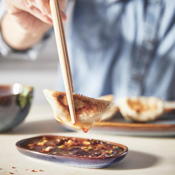 A person holding a dumpling with chopsticks, dipping it into a dark sauce on an ocean-blue, glazed ceramic plate.