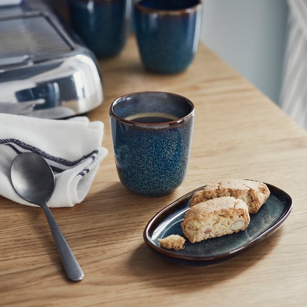 A wooden table has blue ceramic items. A cup of coffee, a plate with biscotti, and a spoon rest near a coffee maker and more cups.