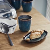 A wooden table has blue ceramic items. A cup of coffee, a plate with biscotti, and a spoon rest near a coffee maker and more cups.