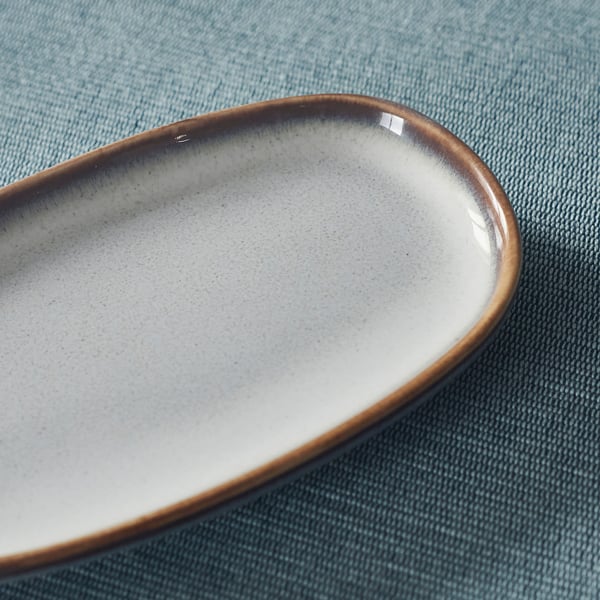 Brown-rimmed elliptical dish with smooth glaze on blue cloth.