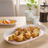 White platter of golden-brown twisted pastries in foreground, alongside mugs, a fruit platter, and a coffee press on wooden table.