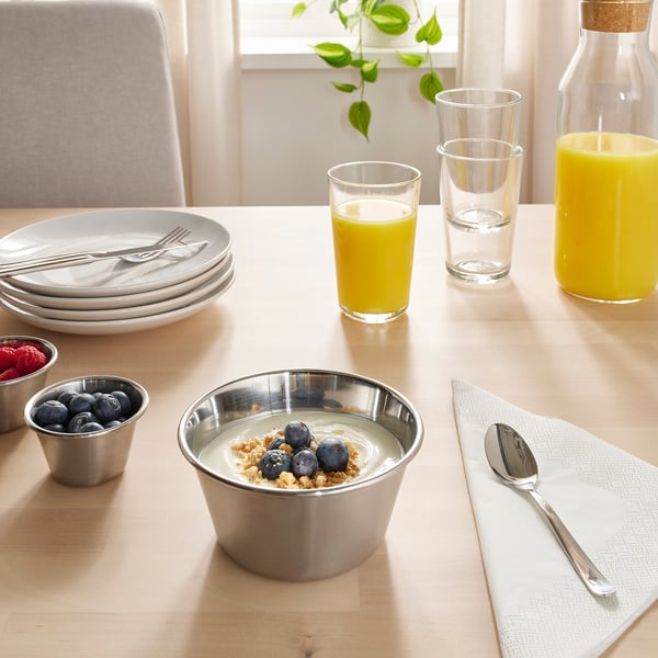 Table set with stainless steel bowl of yogurt, berries, and granola. Glasses of orange juice and water, plates, cutlery, and a plant.