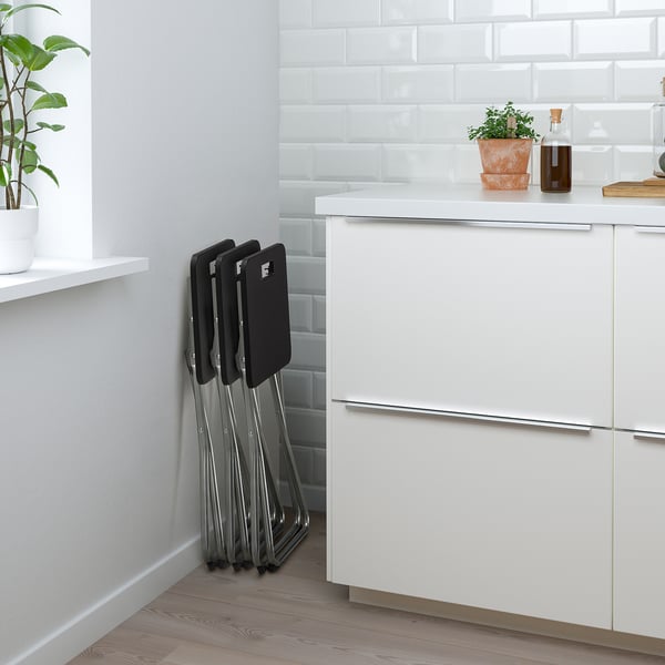 Foldable stackable chairs neatly stored next to a kitchen counter. Chairs are black with metallic legs.