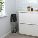 Foldable stackable chairs neatly stored next to a kitchen counter. Chairs are black with metallic legs.