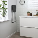 Foldable stool with metal frame hangs on wall, saving space when not in use. Indoor use only.