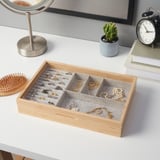 Wooden jewelry box with compartments holds rings, necklaces, and bracelets. Gray felt lining protects items. On a white desk near a brush and clock.