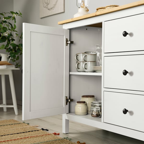 White HEMNES sideboard with open door revealing stored mugs, jars, and plates. Displays household items neatly.