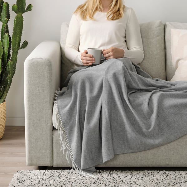 Woman sitting on grey sofa, wrapped in grey fringed throw, holding a mug.