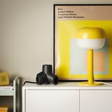 Black HORNFELS 4-way extension cord sits on white furniture, near a yellow lamp. Small, compact, creative design.