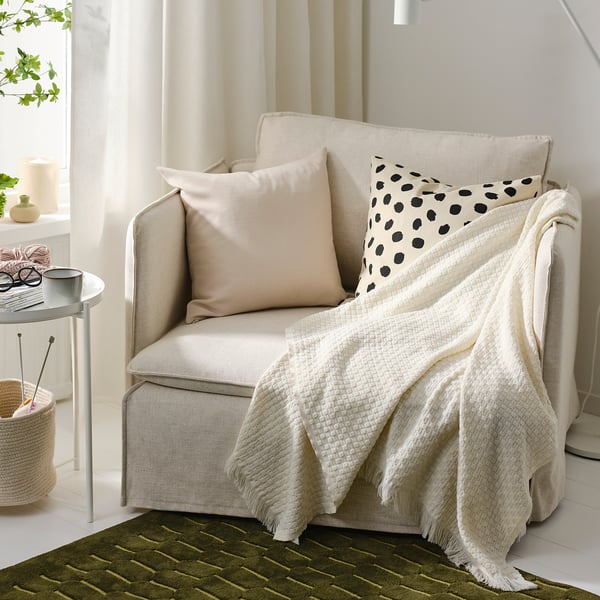 Beige HORNMAL throw blanket on armchair, pillows, side table with mug, knitting, plant, green rug - cozy setting.