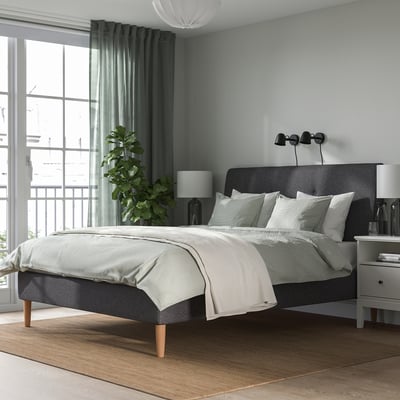 Modern bedroom with upholstered IDANÄS bed, dark grey, sloping headboard, wooden legs. Minimalist design with neutral tones, large window, rug, plant.