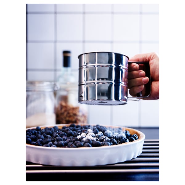 A hand uses a silver flour sifter over a pie filled with blueberries, highlighting baking preparation.