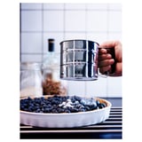 A hand uses a silver flour sifter over a pie filled with blueberries, highlighting baking preparation.
