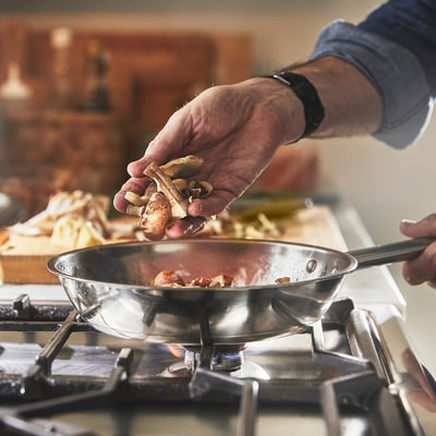 A person is cooking by adding mushrooms to a sizzling pan on a gas stove.