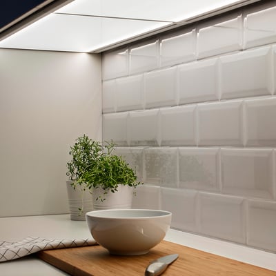 Modern kitchen counter with white bowl, knife, and potted herbs on wooden board near light panel under cabinet.