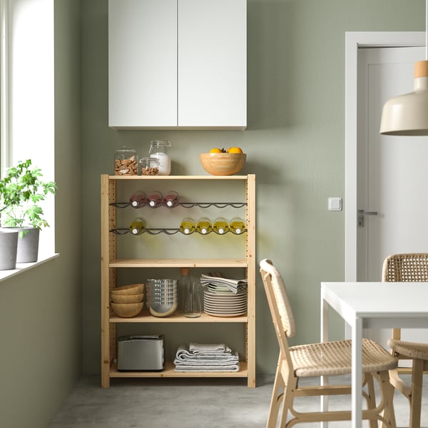 Modern kitchen corner with IVAR pine shelf. Holds jars, plates, and wine. Next to white table and wicker chair. Green wall and white cabinet.