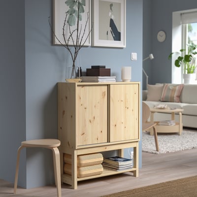 IVAR light wood cabinet stands in blue room with sofa, chair, and plants. Sliding door design, small size against wall.
