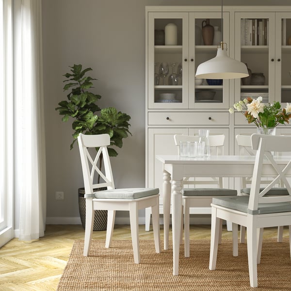 White dining table with JUSTINA chairs, cushioned. Set with glasses and flowers. Minimalistic, cozy room with wood floor, rug, cabinet, and plant.