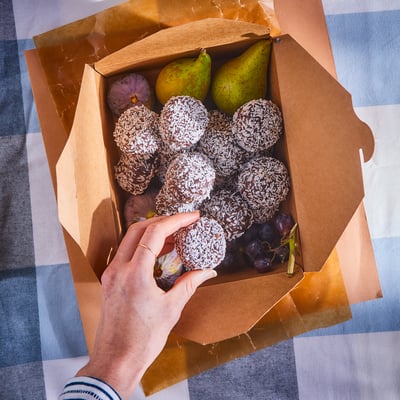 A box of colorful fruits and coconut-rolled chocolate pastries on a striped tablecloth. A hand picks a pastry.
