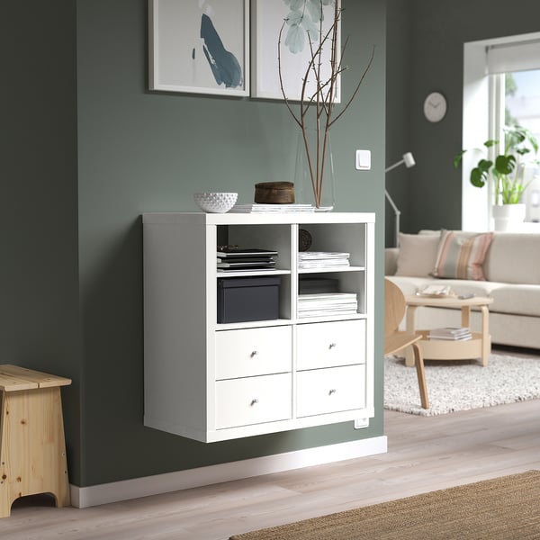 A white KALLAX unit against a green wall with shelves and drawers, displaying various items. A cozy living room setting, noticeable clock and plants.