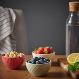 Three colorful bowls filled with cashews, strawberries, and blueberries on a wooden table with a striped cloth, a glass bottle, and a lemon slice.
