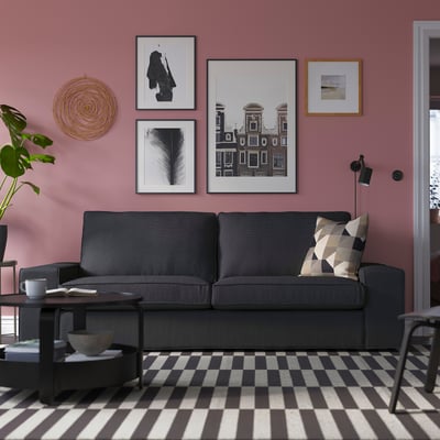 A cosy living room a dark grey KIVIK sofa with a chequered rug, bold art frames against pink walls, and a small side table with decor.